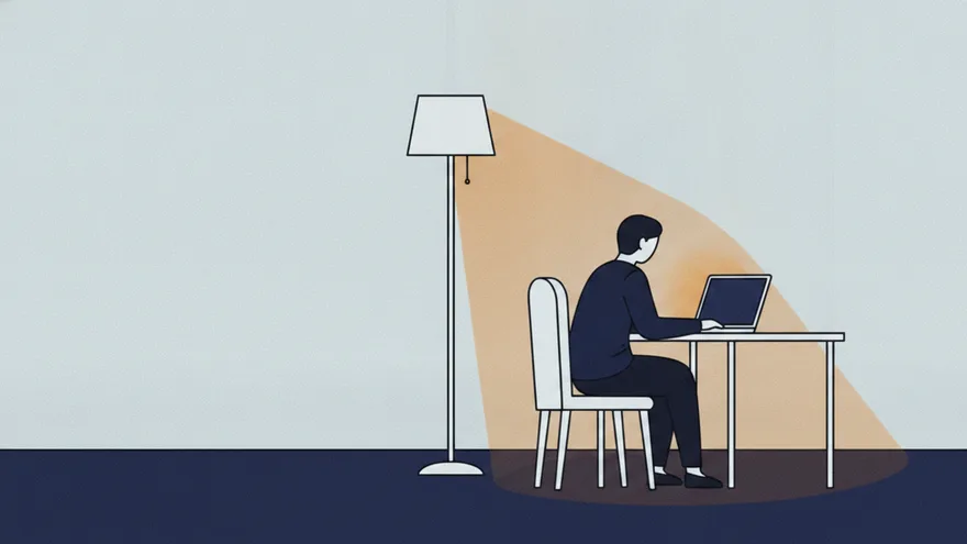 A person alone at a desk in the evening, seen from behind, typing on a laptop whose screen is turned away and blank, lit by a single floor lamp.