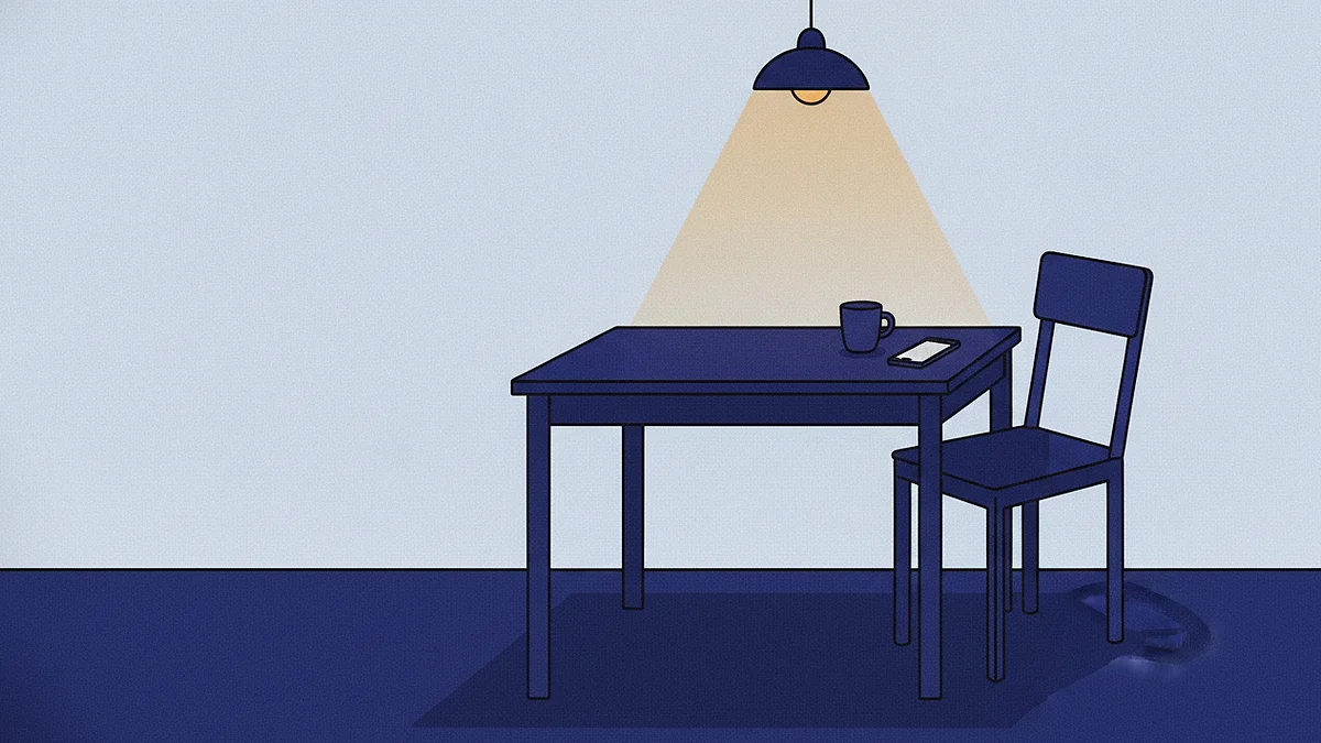 A phone lying flat and dark on a kitchen table beside a cooling cup and a chair pushed back from the table, one lamp lit above.